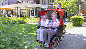 Eine Ehrenamtliche schiebt zwei Seniorinnen in einer Rikscha durch den Garten einer Pflegeeinrichtung.