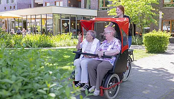 Eine Ehrenamtliche schiebt zwei Seniorinnen in einer Rikscha durch den Garten einer Pflegeeinrichtung.