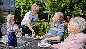 Eine Ehrenamtliche serviert im Garten einer Senioreneinrichtung Kaffee und Kuchen an einem Tisch.