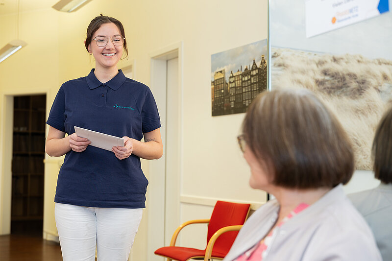 Mitarbeiterin steht in einem Praxisraum und hält ein Formular in der Hand, während eine sitzende Patientin ihr zuhört.