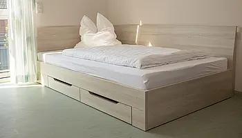 Einzelbett mit Bettkasten in einem hellen Apartmentzimmer mit Tageslicht.