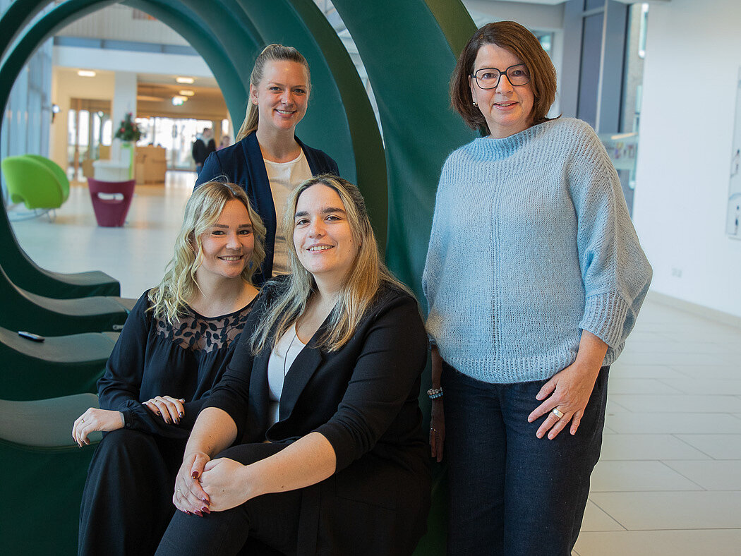 Zwei Ausbildungskoordinatorinnen stehen in der Magistrale, zwei sitzen daavor: Alle lächeln freundlich und zugewandt.