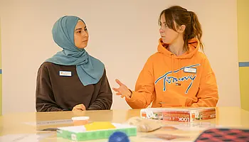 Zwei junge Frauen sitzen an einem Tisch und tauschen sich über Aufgaben und Erfahrungen in einem sozialen Arbeitsbereich aus.