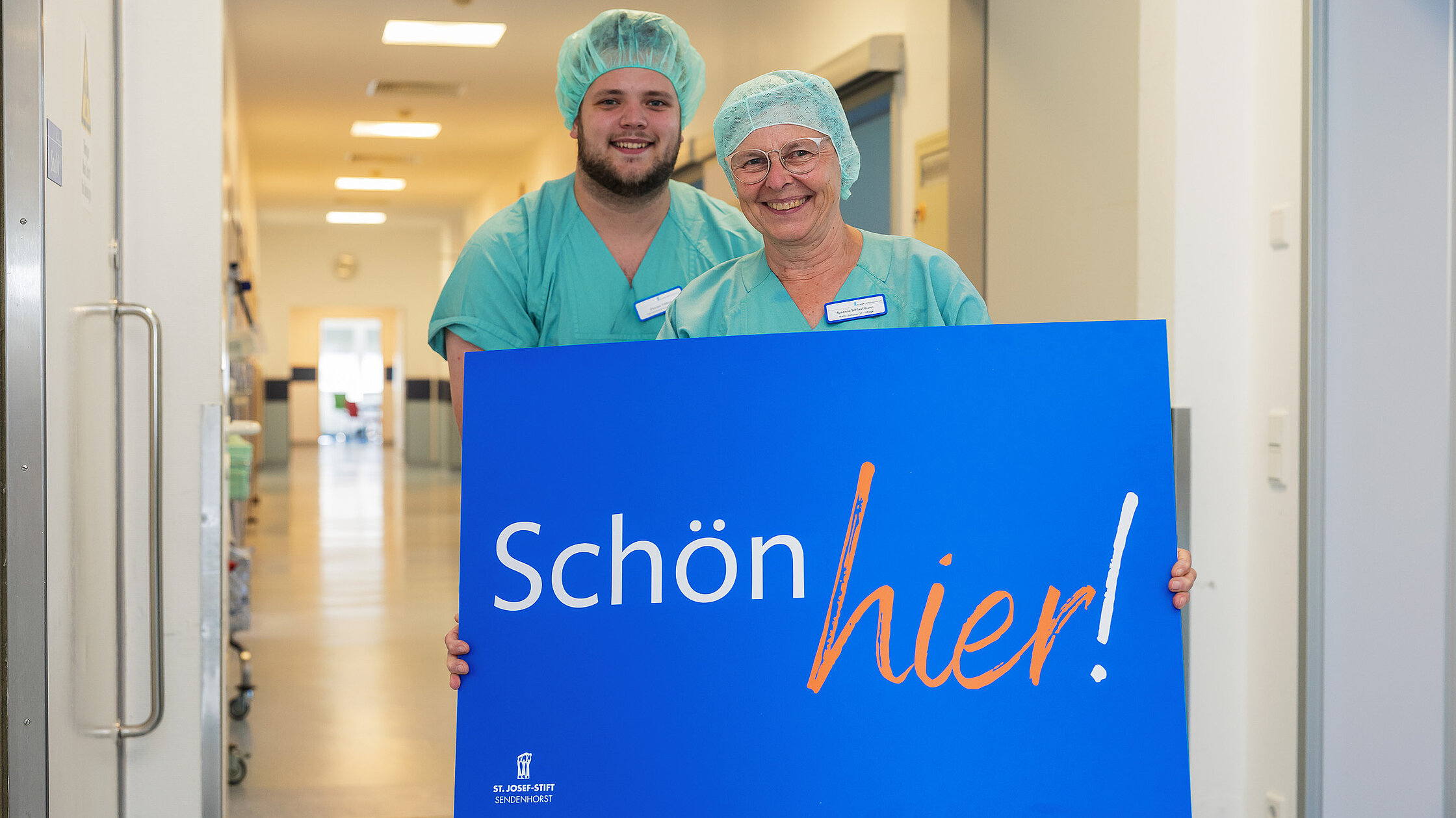  Zwei Mitarbeitende in OP-Kleidung stehen auf einem Flur und halten ein Schild mit der Aufschrift „Schön hier!“.