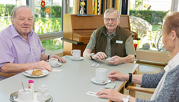 Drei ältere Menschen sitzen an einem Tisch, trinken Kaffee und spielen gemeinsam Karten.