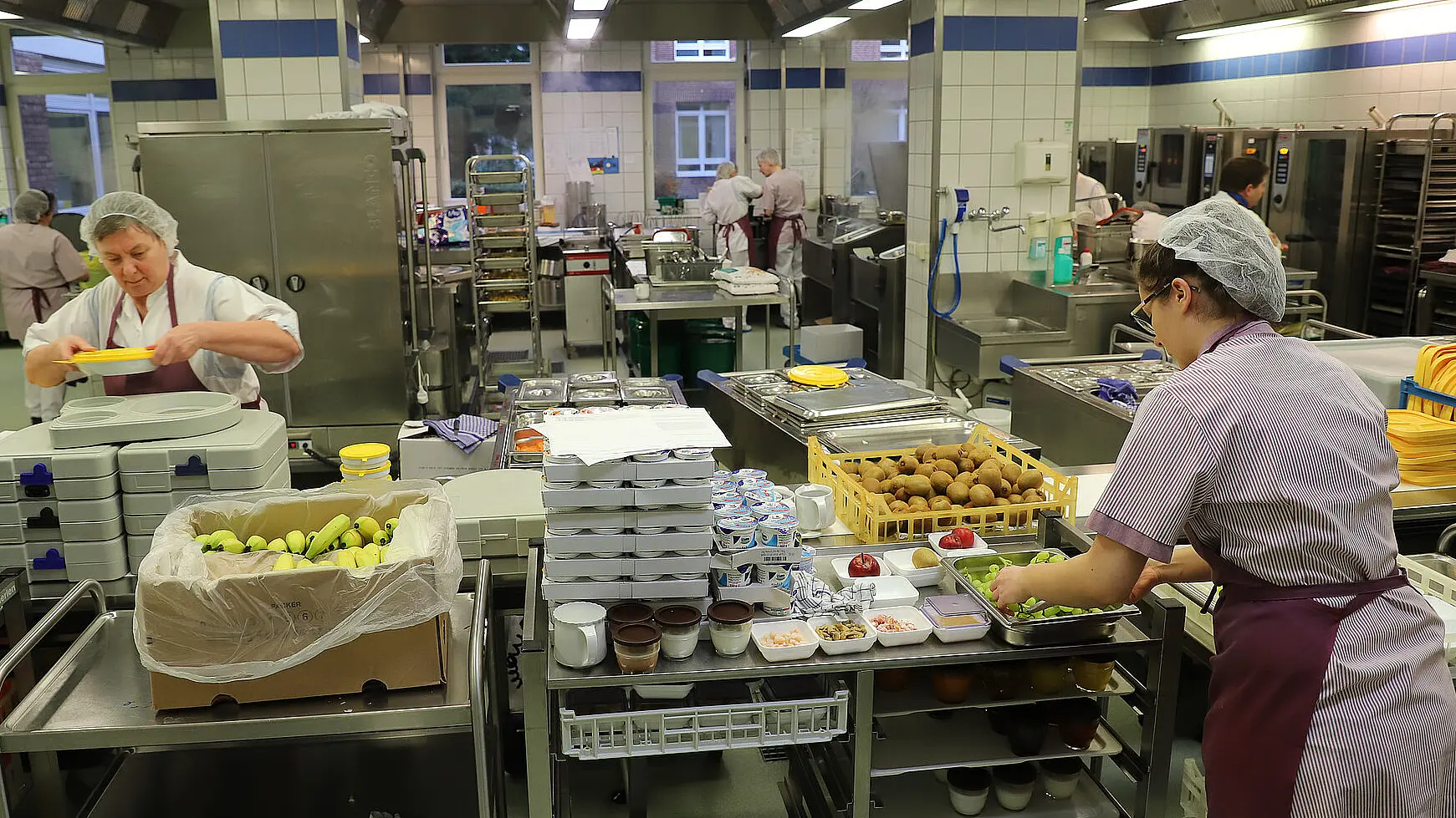 Mehrere Mitarbeitende bereiten in einer Großküche Speisen vor und richten Arbeitsmaterialien an Edelstahlwagen.