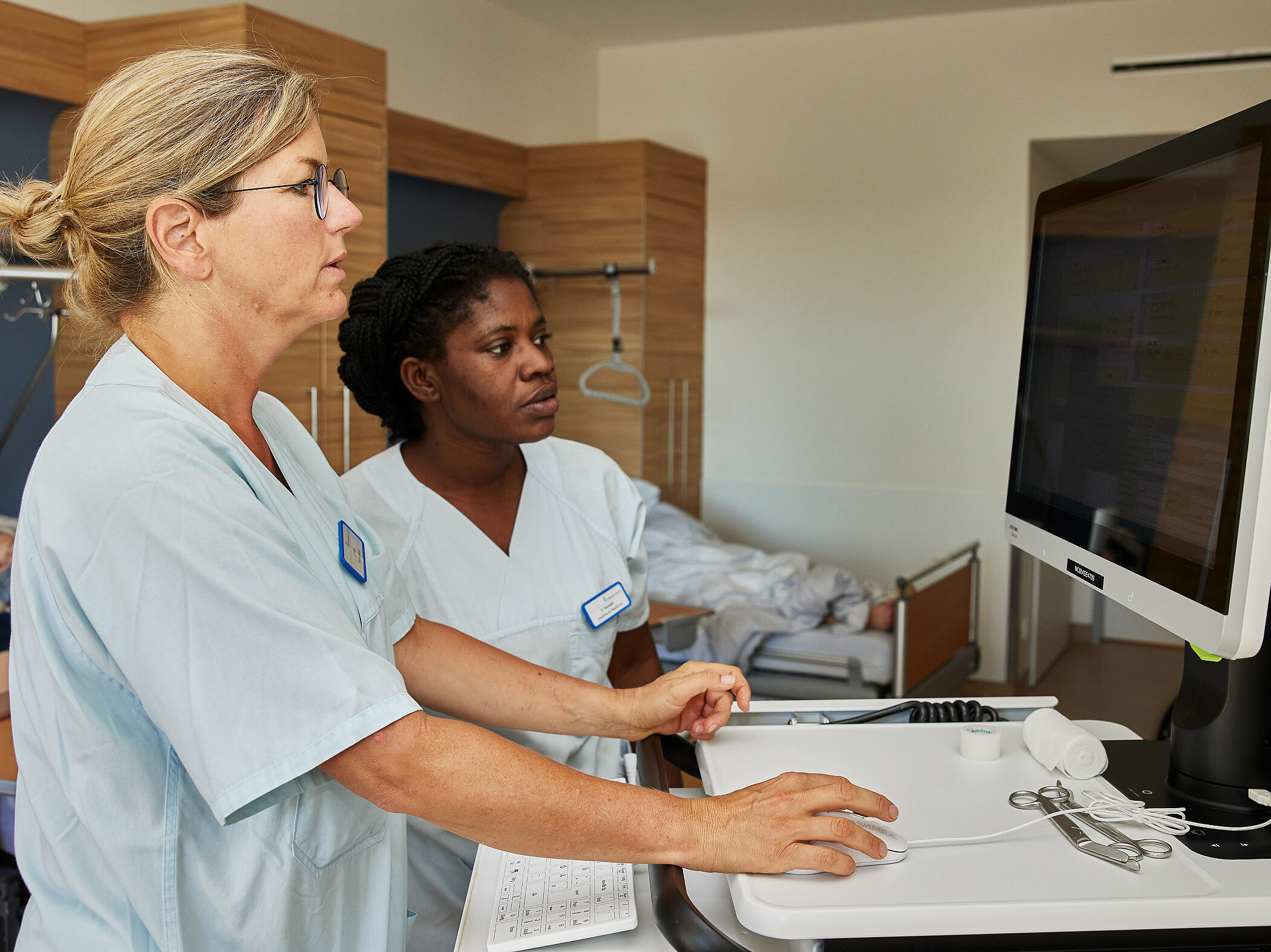 Zwei Pflegekräfte stehen gemeinsam an einem mobilen Computerwagen und arbeiten am Bildschirm.