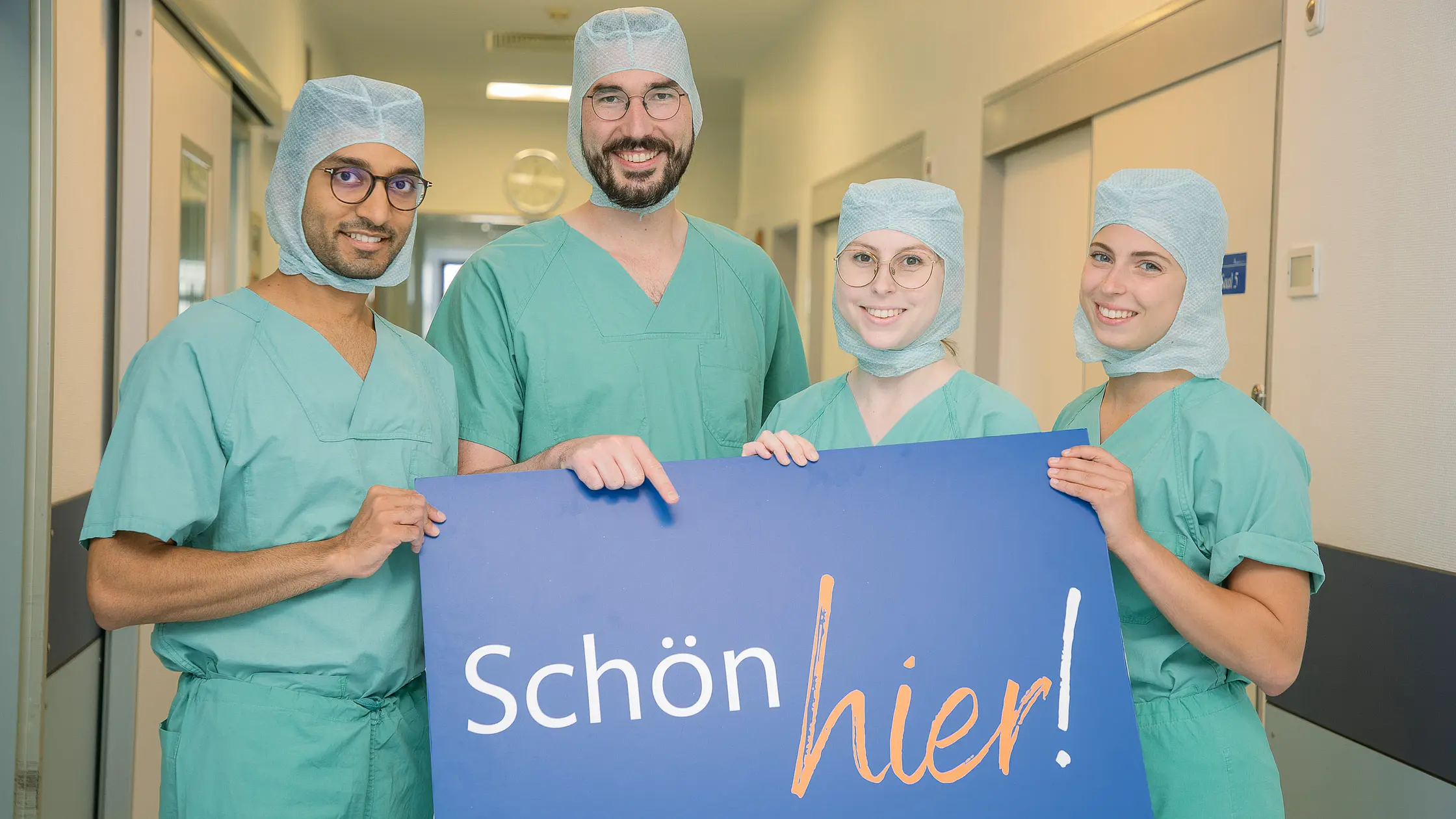 Vier Personen in grüner OP-Kleidung stehen auf einem Klinikflur und halten ein Schild mit der Aufschrift „Schön hier!“.