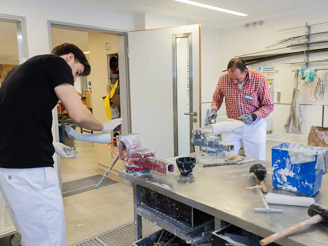 Zwei Mitarbeiter bearbeiten gleichzeitig Gipsleistenmodelle an einer zentralen Werkbank mit Werkzeugen und Materialbehältern.