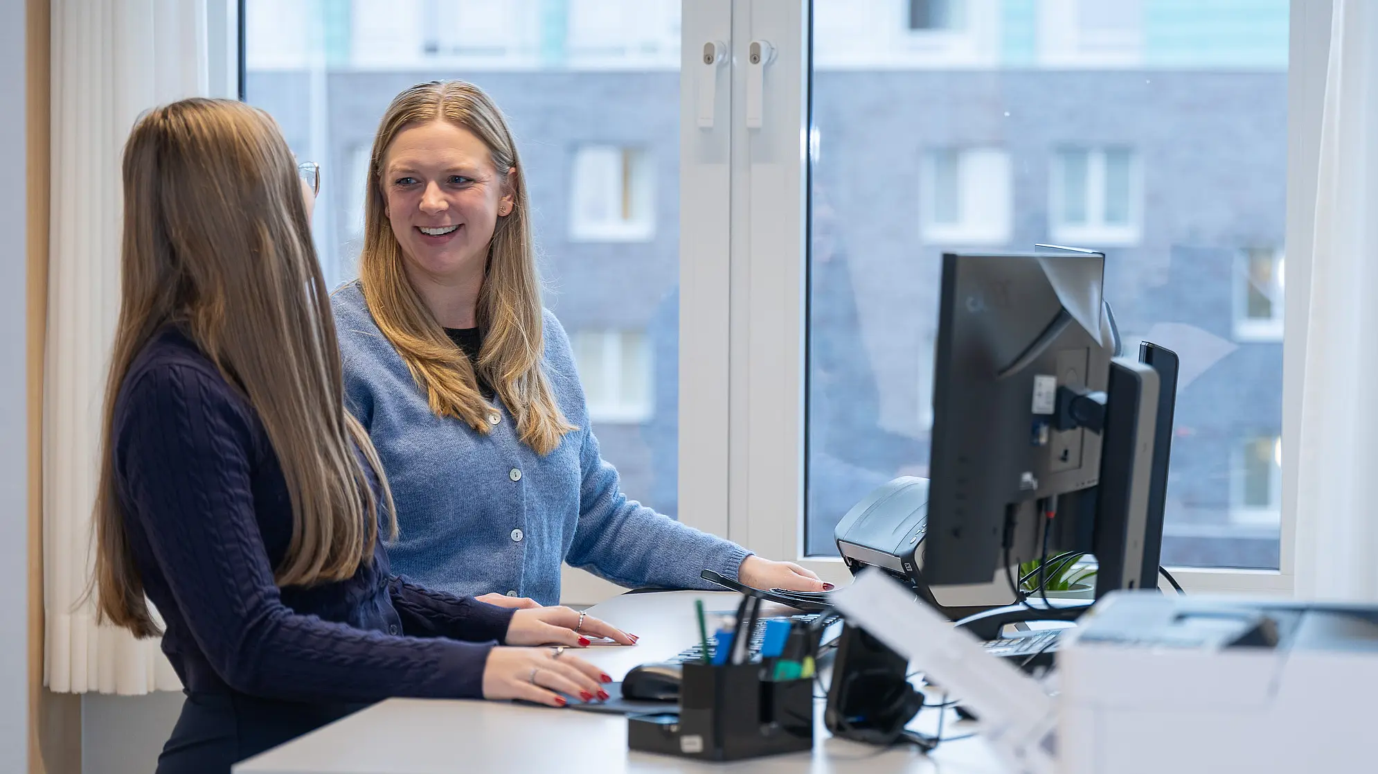 Eine Auszubildende und ihre Praxisanleitung stehen in einem Büro an einem Schreibtisch und unterhalten sich vor einem Computerarbeitsplatz.