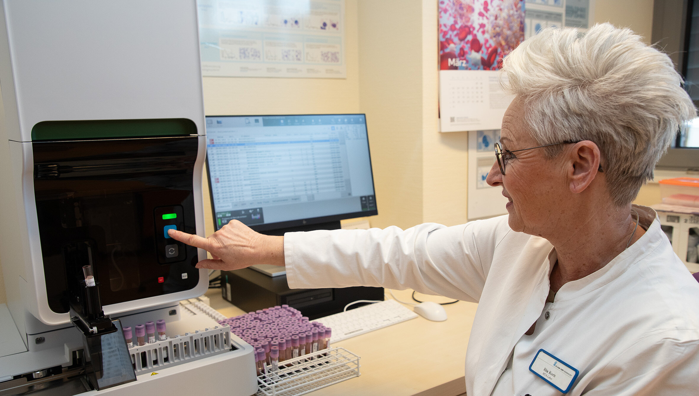 Eine Medizinisch Technische Assistentin im Labor bedient ein Analysegerät zur Auswertung von Blutproben, im Hintergrund ein Computer mit Patient:innendaten.