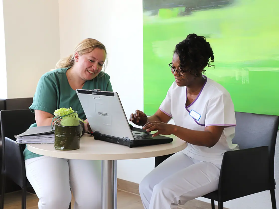 Eine Auszubildende sitzt mit einer Praxisanleitung am Tisch und hat ein Laptop vor sich, beide schauen darauf.