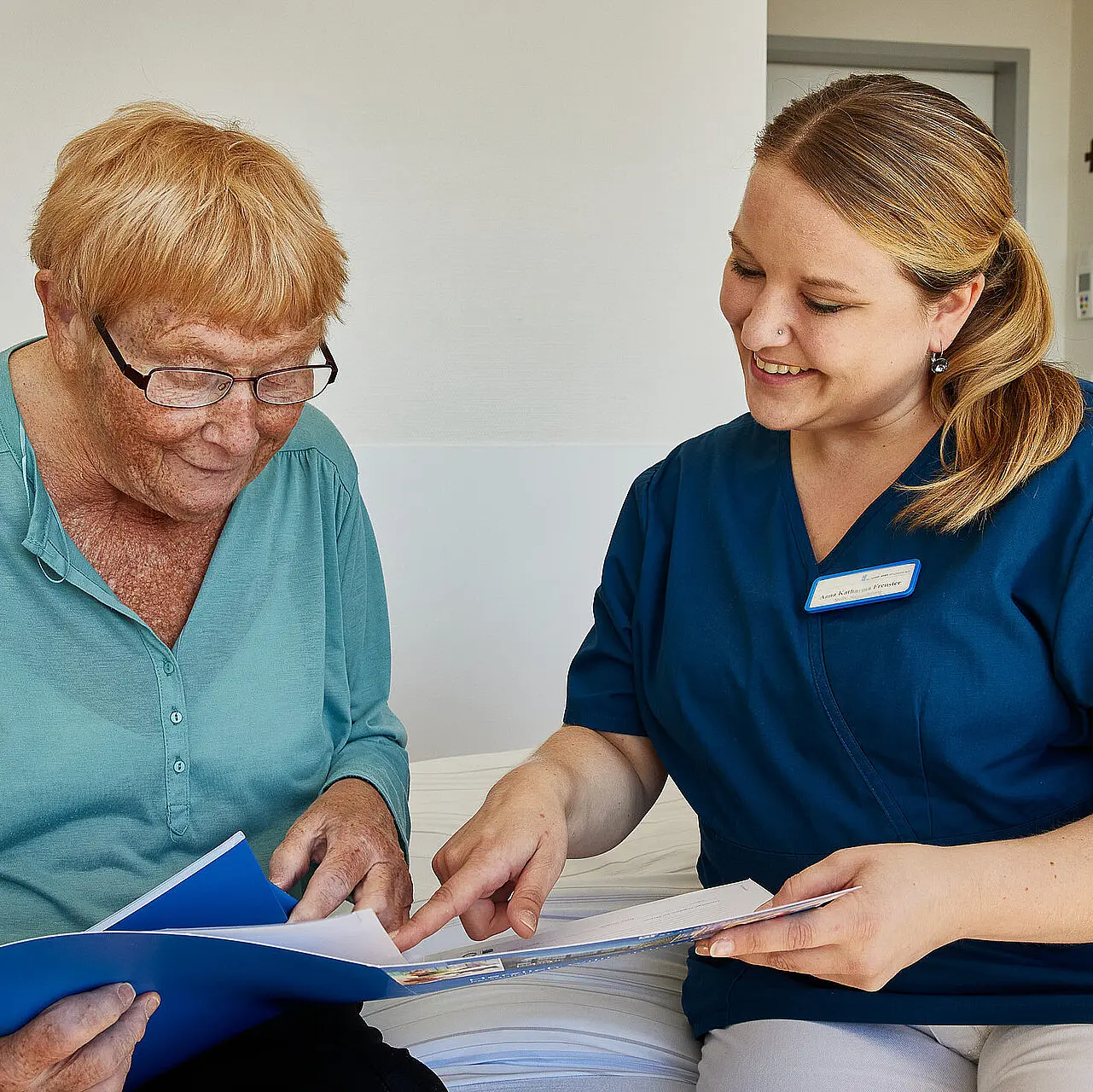 Eine Pflegekraft erklärt einer älteren Person Unterlagen, beide sitzen gemeinsam auf einem Bett.