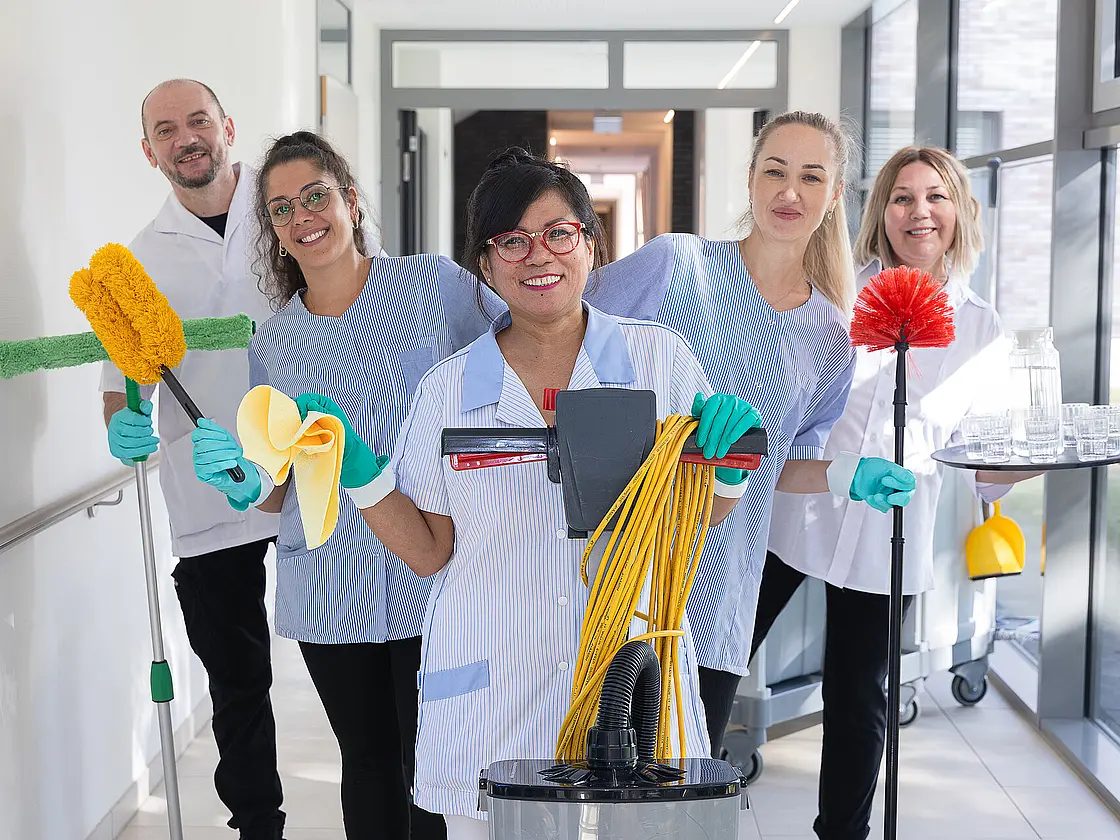 Fünf Mitarbeitende aus der Reinigung stehen mit Reinigungsutensilien wie Wischmopp, Tuch und Tablett in einem Flur und lächeln in die Kamera.