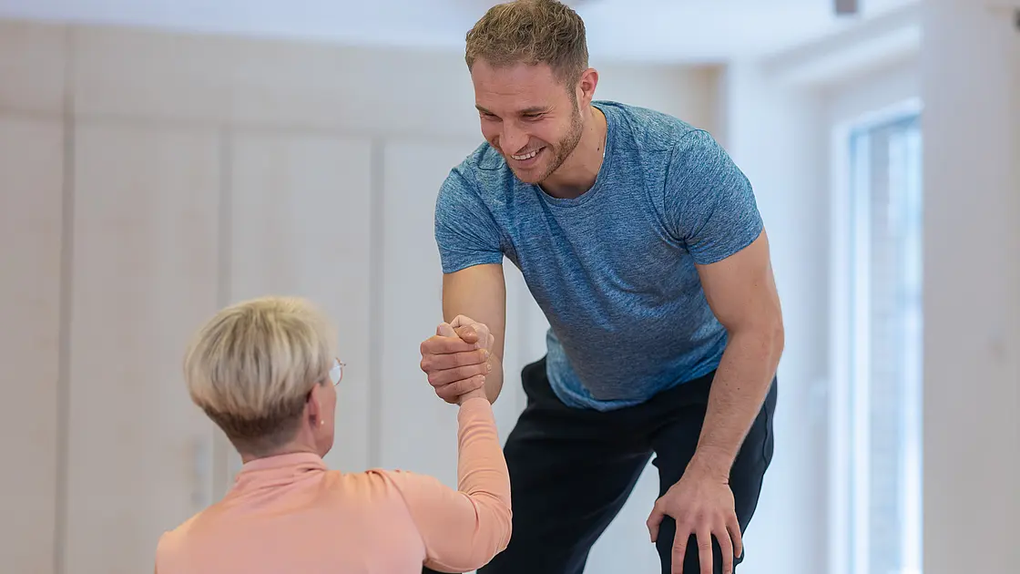 Ein Therapeut unterstützt eine Patientin beim Aufstehen und stabilisiert sie mit einem Handgriff.