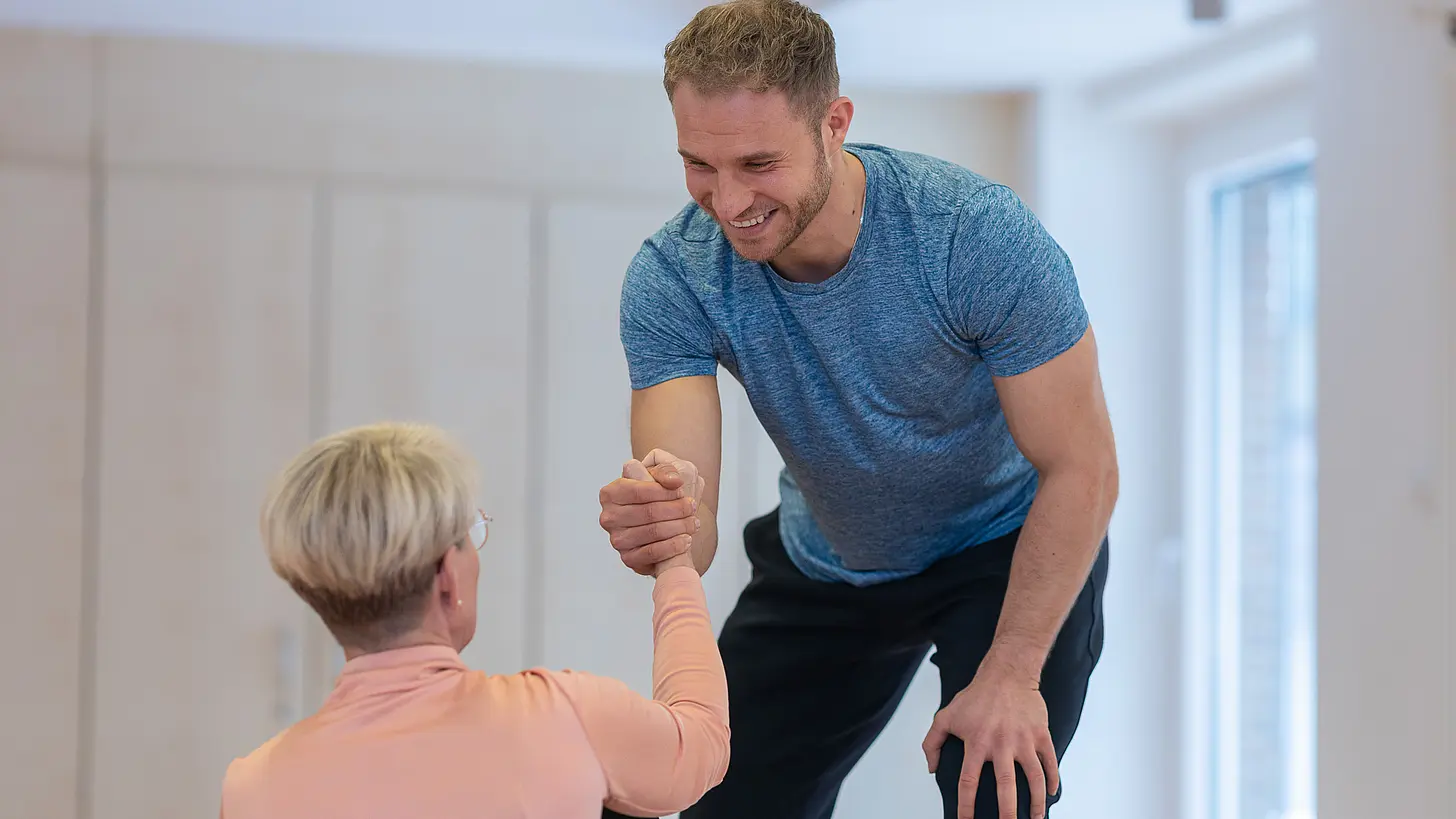 Ein Therapeut unterstützt eine Patientin beim Aufstehen und stabilisiert sie mit einem Handgriff.