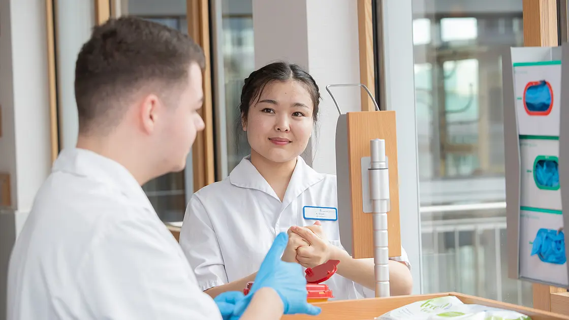 Zwei Auszubildende stehen an einem Desinfektionsspender im Stationsflur und führen eine Händedesinfektion durch.