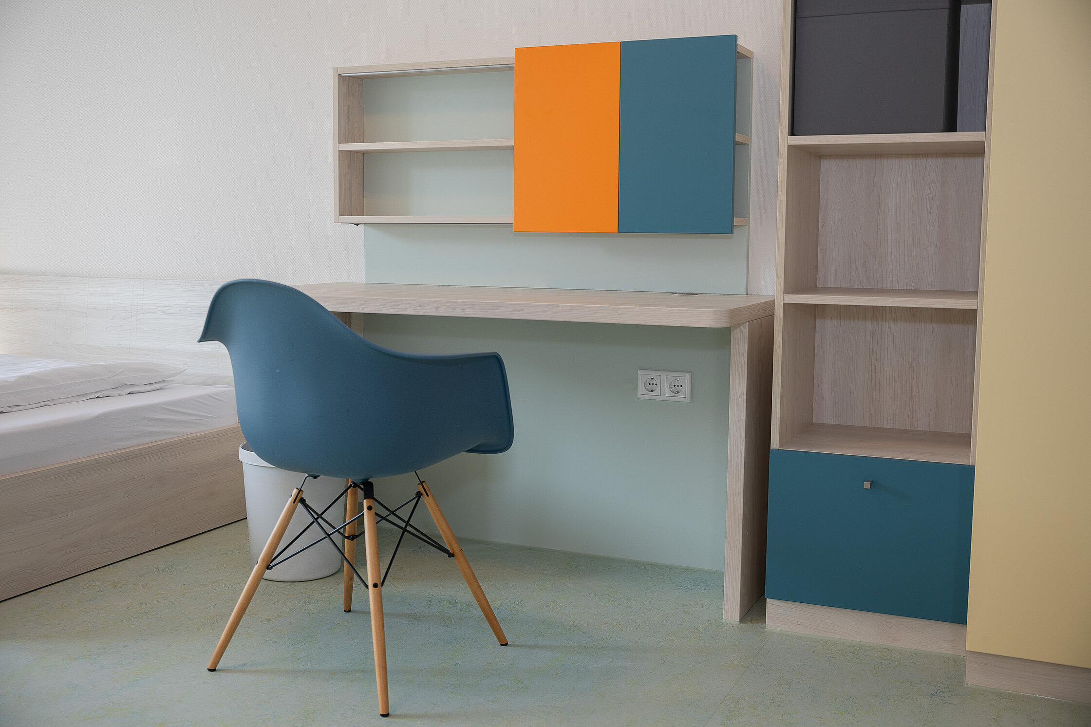 Zu sehen ist die Einrichtung im Apartmenthaus. Ein Stuhl steht vor einem Schreibtisch, daneben sind ein Bett und ein Schrank.