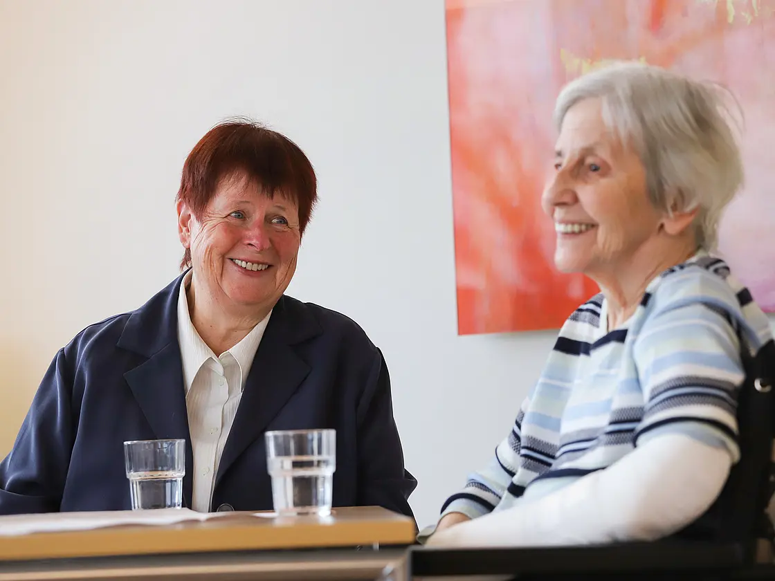 Zwei Frauen sitzen an einem Tisch im Gespräch; vor ihnen stehen Gläser.