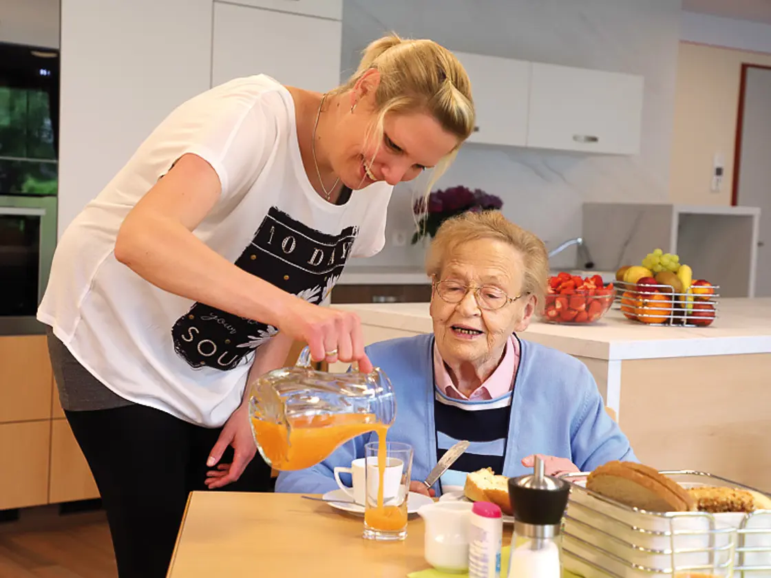 Eine Mitarbeiterin schenkt einer älteren Person am Tisch ein Getränk ein; Frühstücksgeschirr steht bereit.