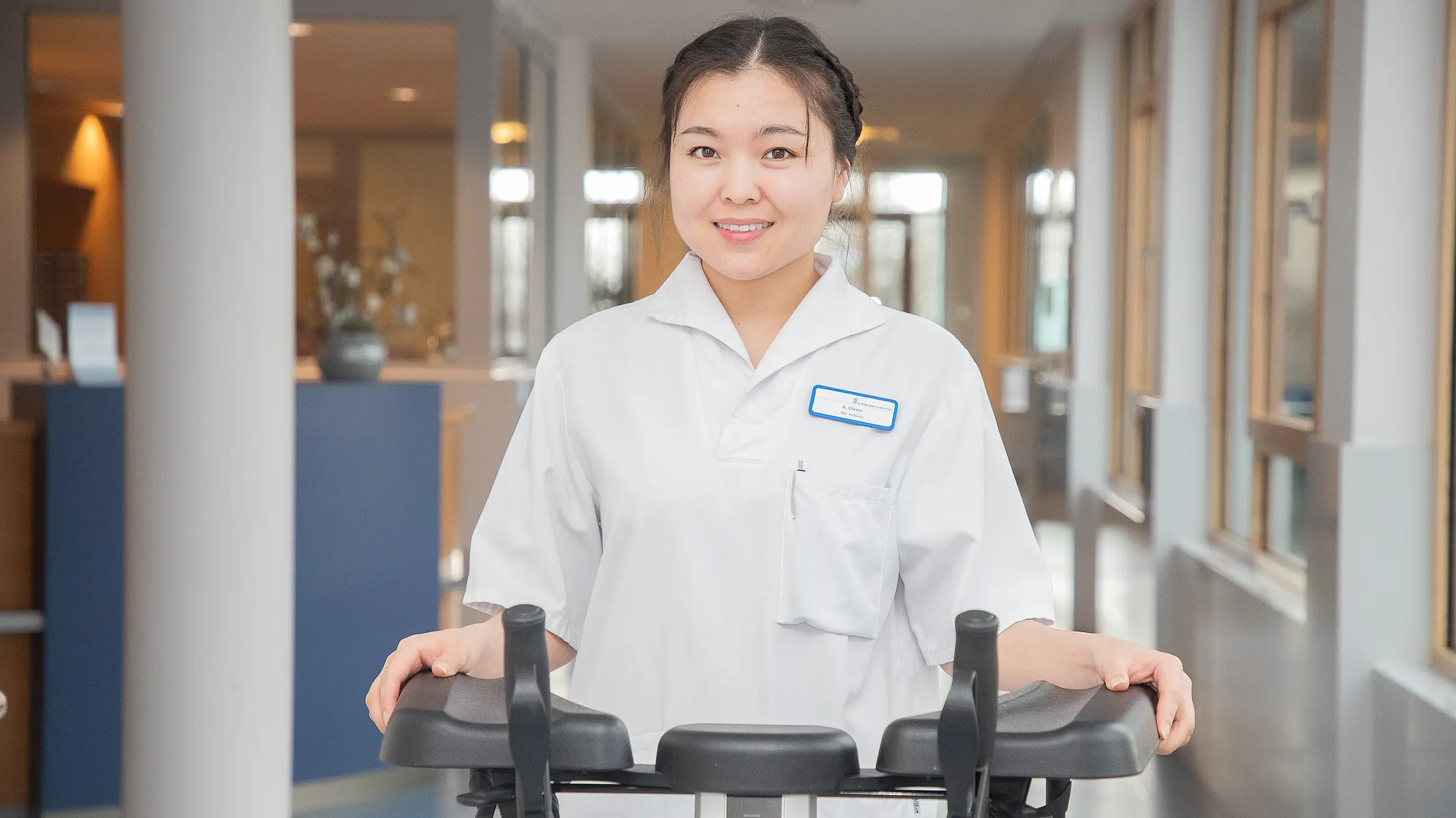 Eine Auszubildende führt einen Gehwagen durch einen Flur mit Fensterfront und Behandlungsräumen.