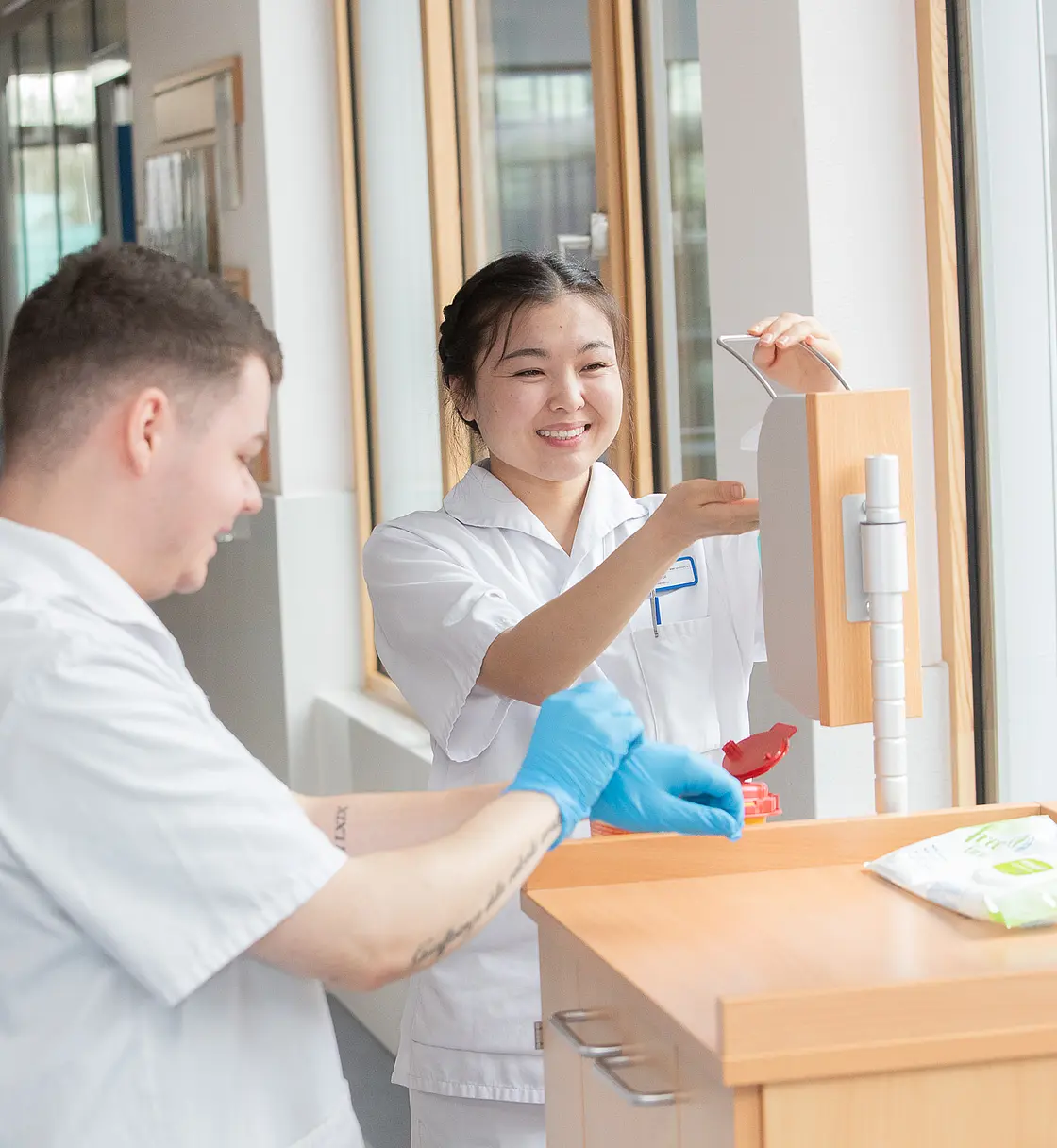 Eine Auszubildende bedient einen Desinfektionsspender, während ein Kollege mit Handschuhen daneben steht.