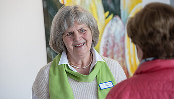 Ehrenamtliche Mitarbeiterin mit grünem Halstuch spricht freundlich mit einer Besucherin im Klinikgebäude.