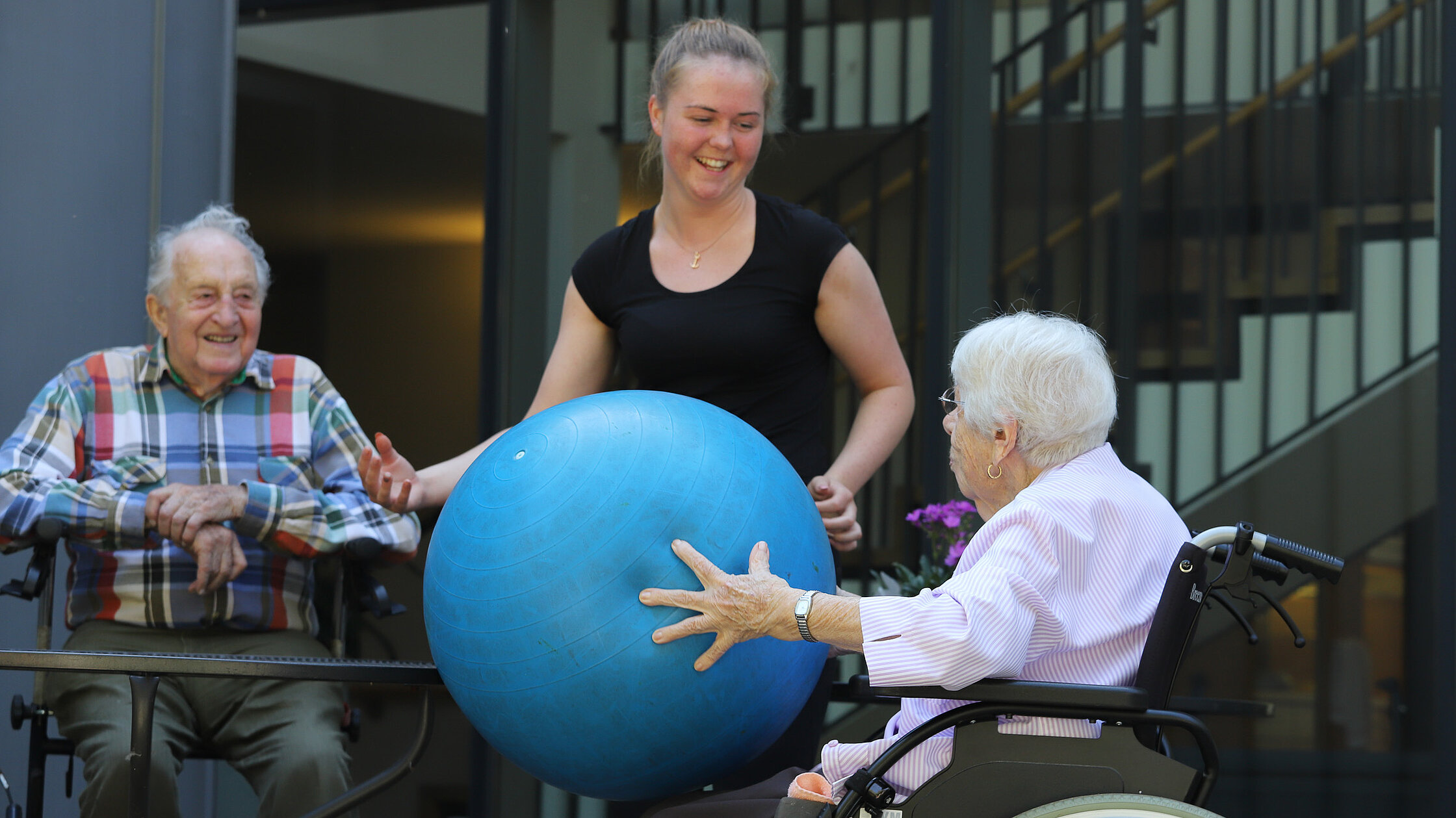 Junge Frau unterstützt zwei Senior:innen im Rollstuhl bei einer Bewegungsübung mit einem großen Gymnastikball im Innenhof einer Einrichtung.