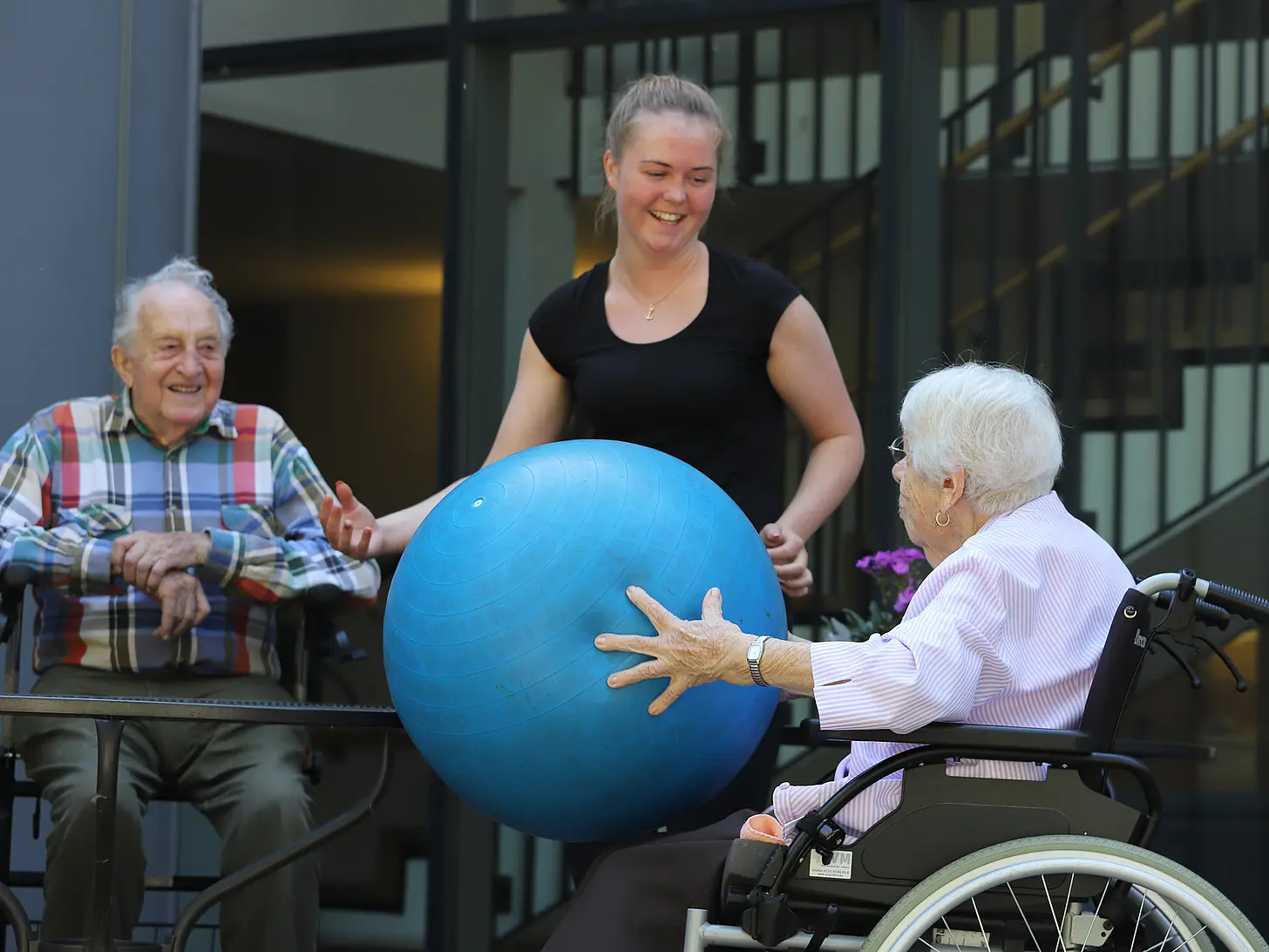 Junge Frau unterstützt zwei Senior:innen im Rollstuhl bei einer Bewegungsübung mit einem großen Gymnastikball im Innenhof einer Einrichtung.