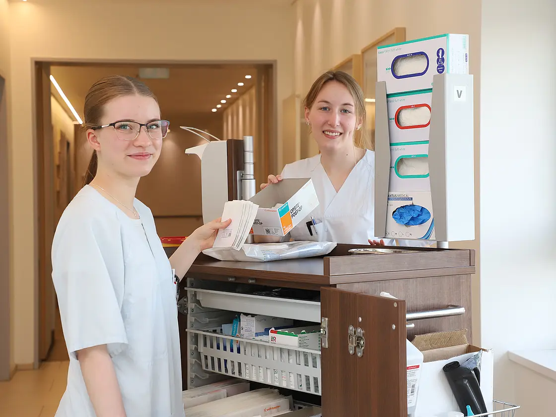Zwei FOSlerinnen stehen an einem Pflegewagen im Krankenhausflur und bereiten gemeinsam Materialien vor.