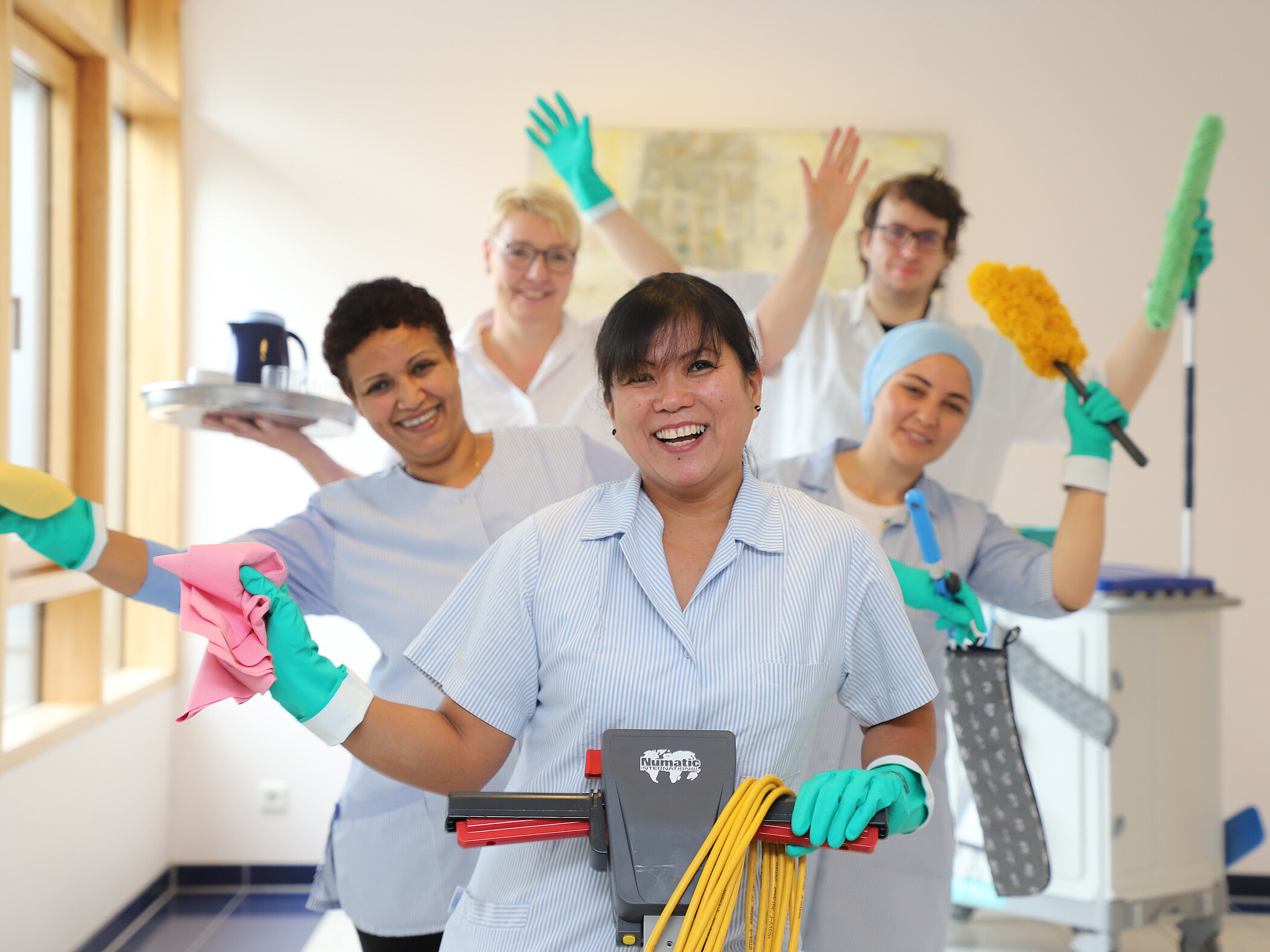 Fünf Mitarbeitende aus der Reinigung stehen mit Reinigungsutensilien wie Wischmopp, Tuch und Tablett in einem Flur und lächeln in die Kamera.