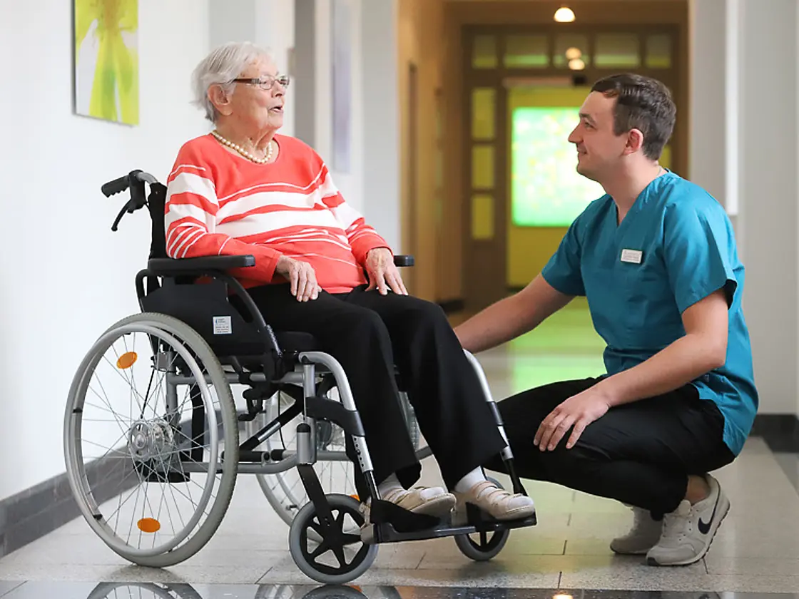 Auszubildender Pflegefachmann spricht mit einer älteren Frau im Rollstuhl auf einem Klinikflur.