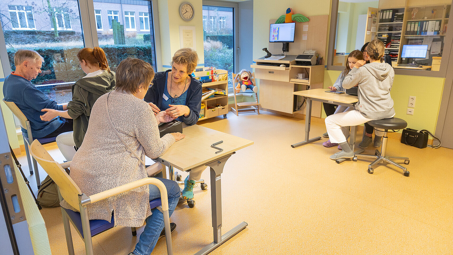 Großer Therapieraum mit mehreren Arbeitsplätzen: Eine Therapeutin begleitet eine erwachsene Patientin bei einer feinmotorischen Übung, während im selben Raum weitere Erwachsene und ein Kind an separaten Tischen therapeutische Aufgaben bearbeiten.
