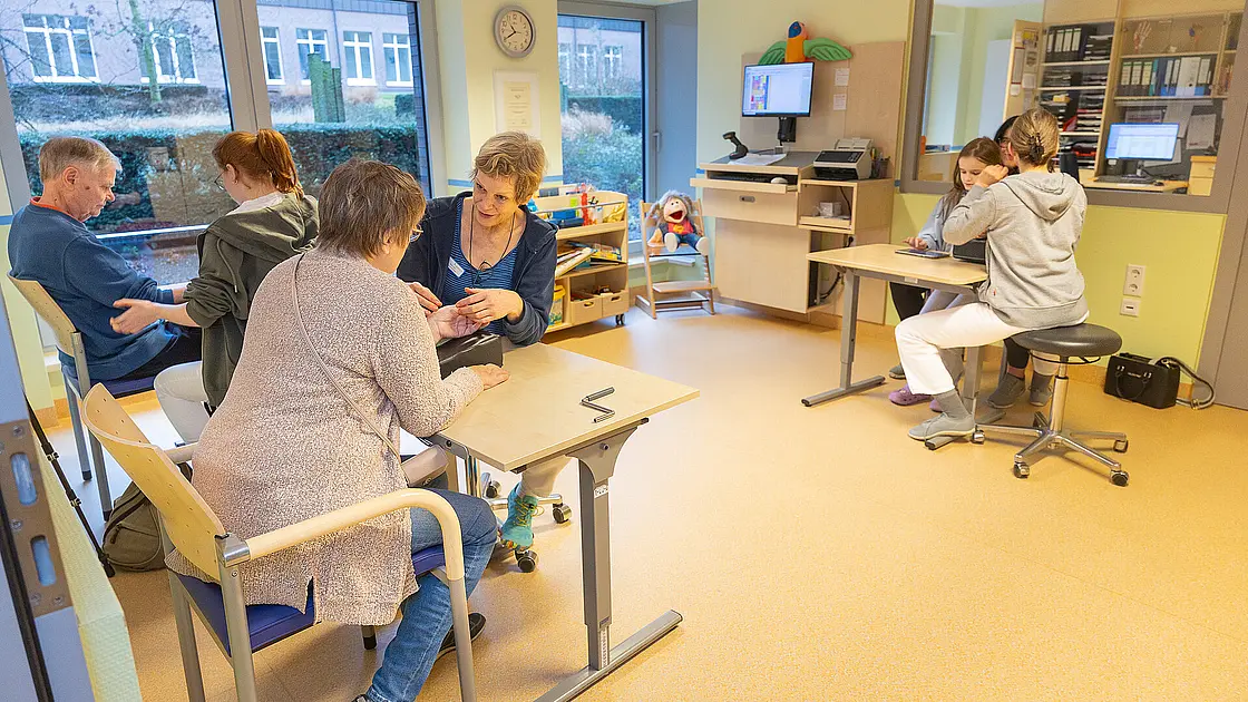 Großer Therapieraum mit mehreren Arbeitsplätzen: Eine Therapeutin begleitet eine erwachsene Patientin bei einer feinmotorischen Übung, während im selben Raum weitere Erwachsene und ein Kind an separaten Tischen therapeutische Aufgaben bearbeiten.