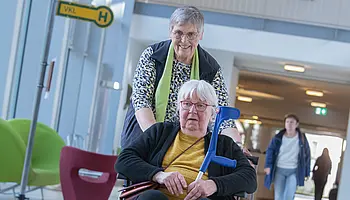 Ehrenamtliche schiebt eine Patientin im Rollstuhl durch den hellen Eingangsbereich der Klinik.