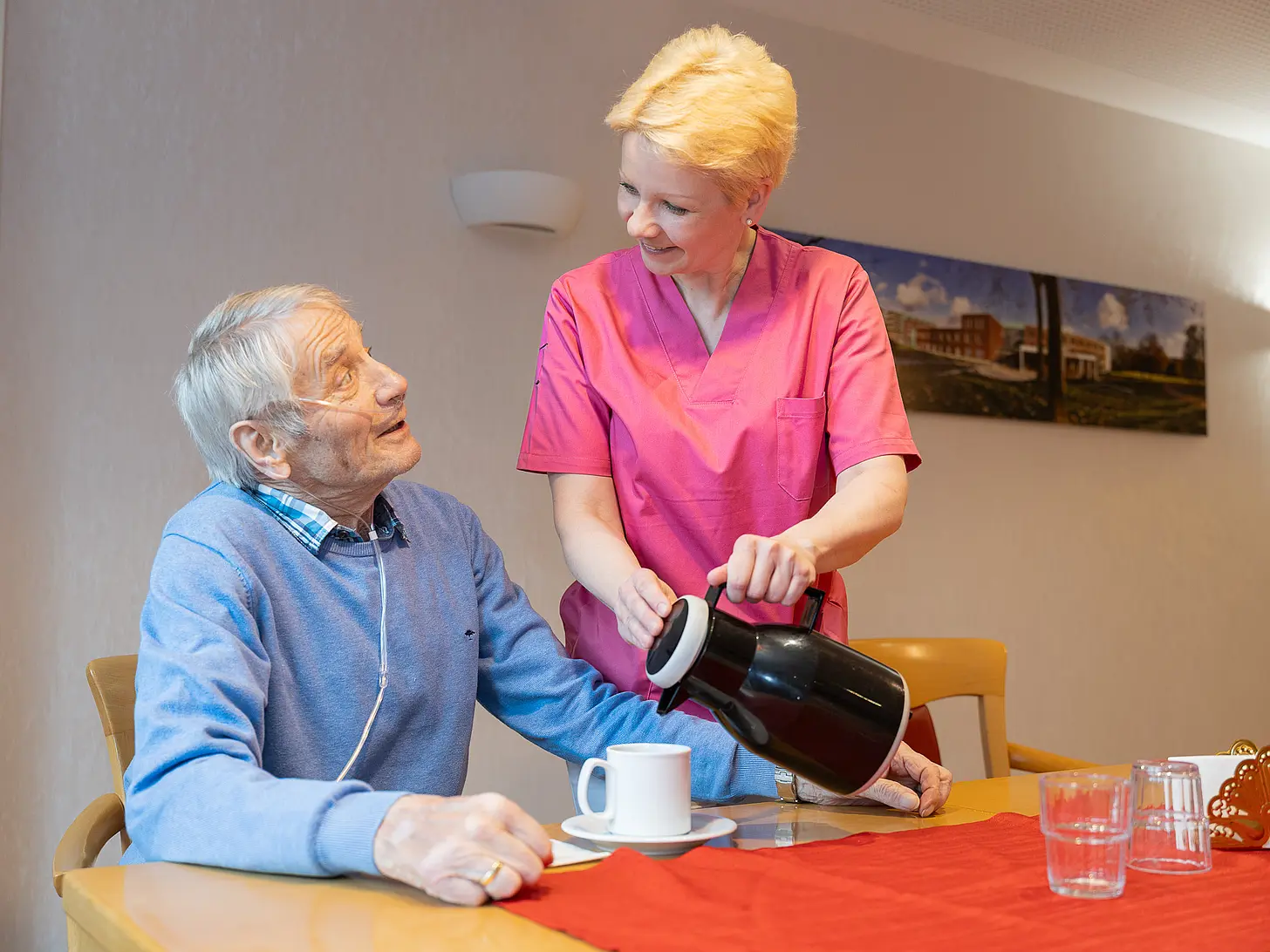 Ein älterer Mann sitzt am Tisch, während eine Pflegefachassistenz ihm Kaffee einschenkt. Beide stehen im Blickkontakt.