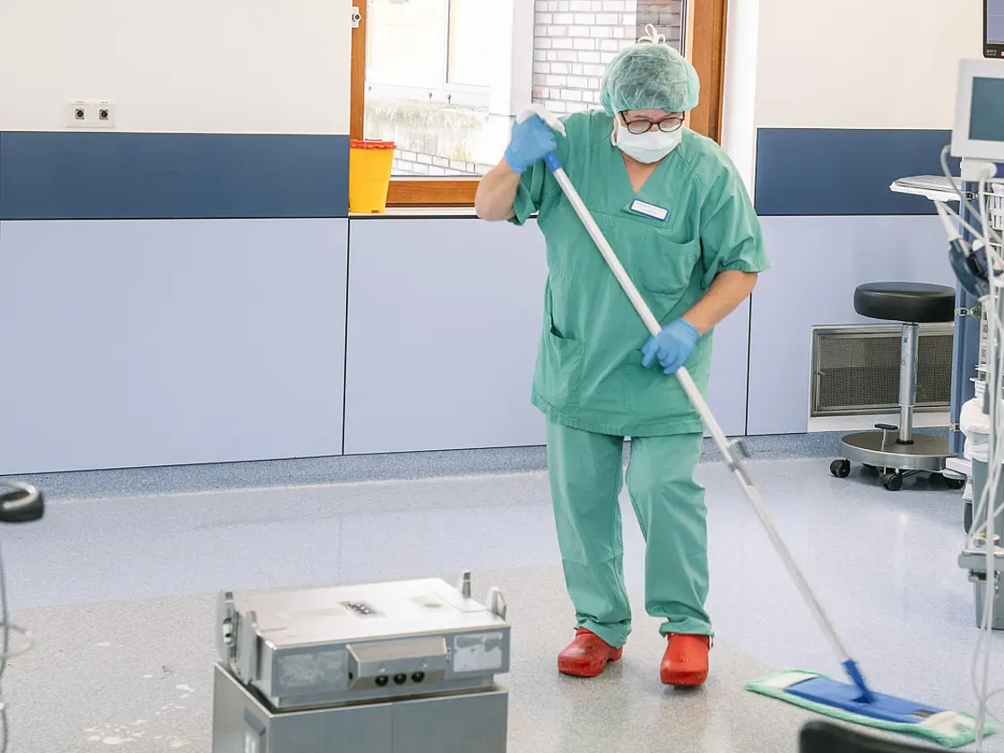 Eine Mitarbeiterin reinigt mit einem Wischsystem den Boden eines Operationsraums; medizinische Geräte und OP-Leuchten sind sichtbar.