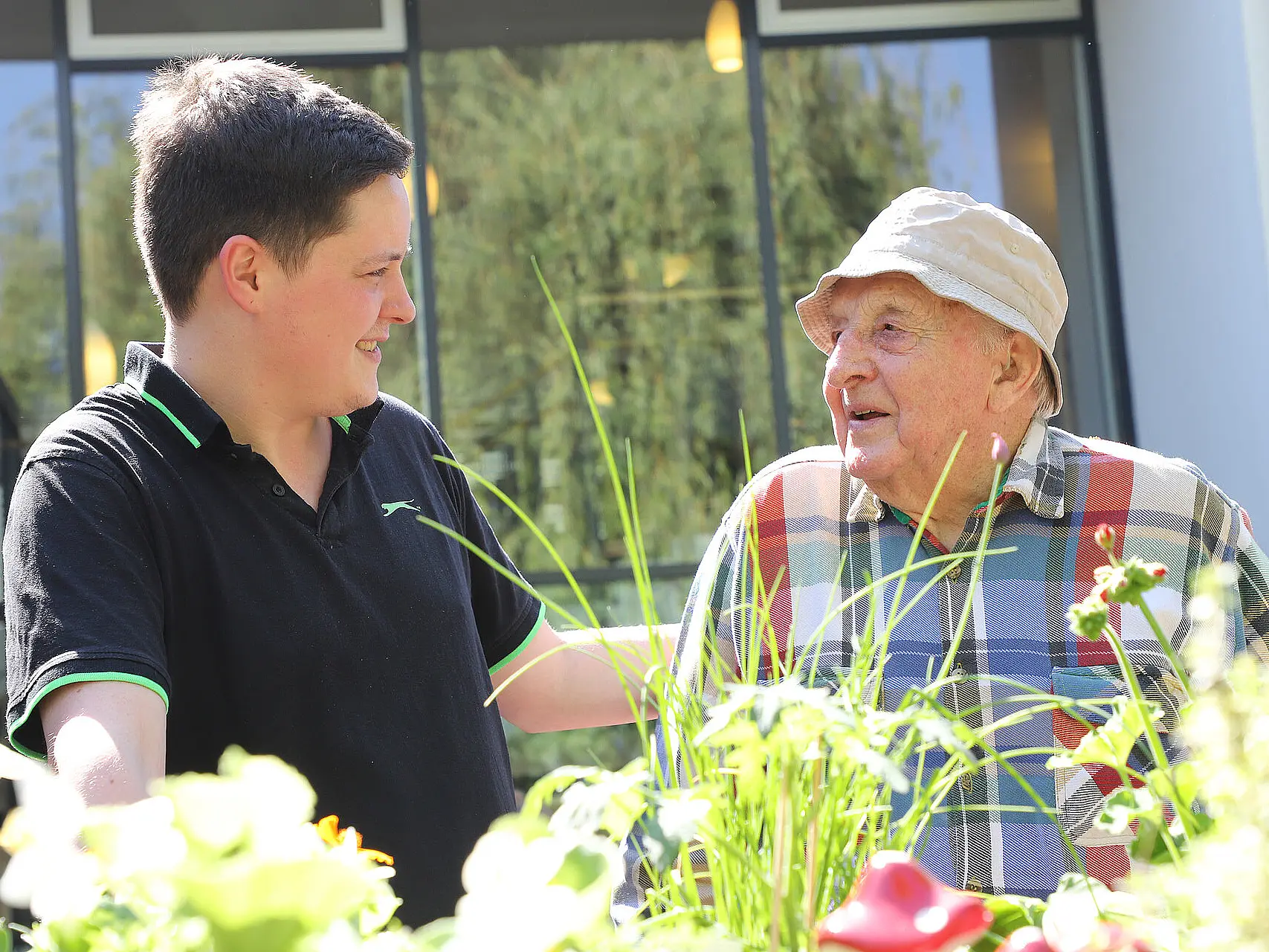 Junger Mann unterhält sich im Garten einer Pflegeeinrichtung freundlich mit einem älteren Mann, umgeben von Blumen und Grünflächen.