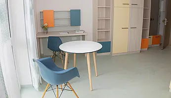 Apartmentzimmer mit rundem Tisch, zwei Stühlen, Schreibtisch und Kleiderschrank.