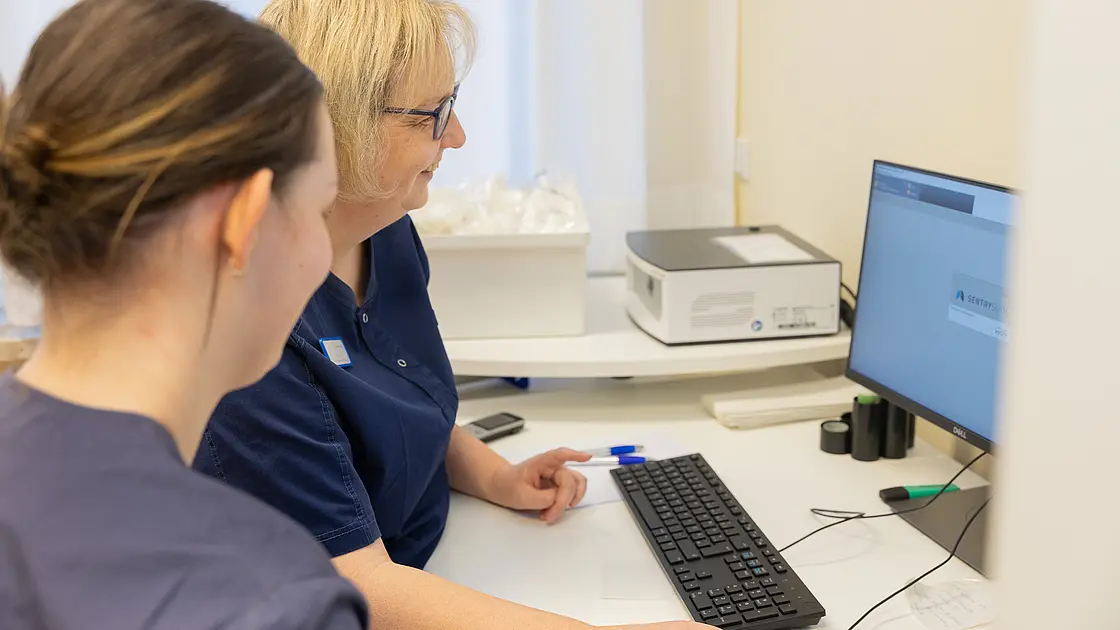 Zwei Mitarbeitende in medizinischer Arbeitskleidung sitzen vor einem Computer und betrachten gemeinsam den Bildschirm.