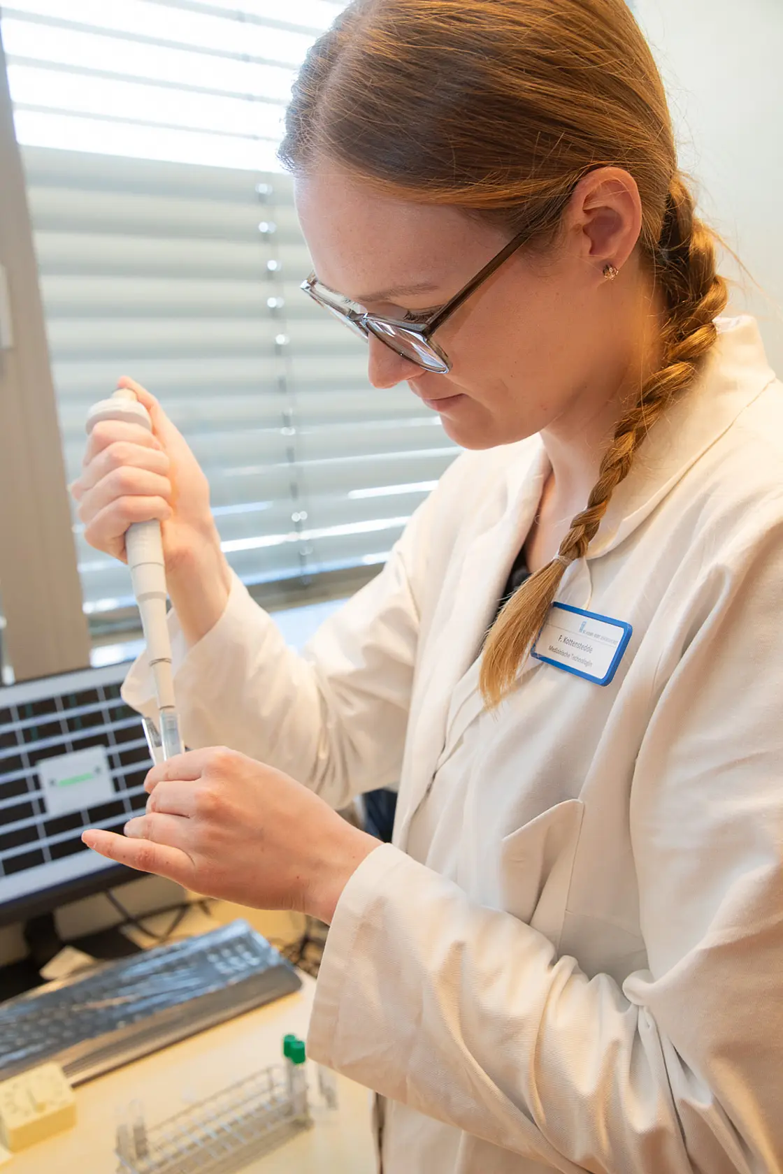 Labor-Mitarbeiterin pipettiert eine Flüssigkeit in ein Probengefäß, im Hintergrund ein Computerarbeitsplatz im Diagnostikzentrum.