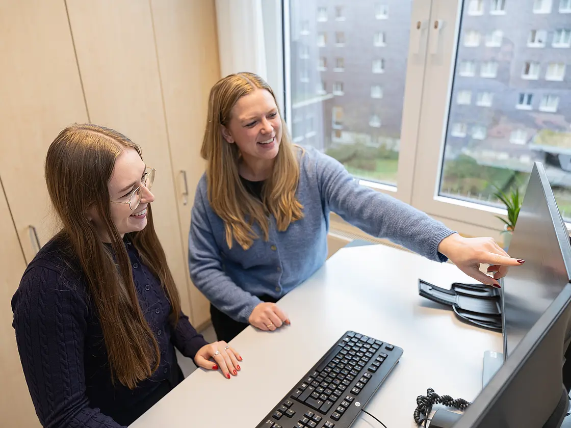 Eine Auszuibildende und ihre Praxisanleitung sitzen an einem Schreibtisch und betrachten gemeinsam einen Computerbildschirm, die Praxisanleitung zeigt erklärend auf den Monitor.