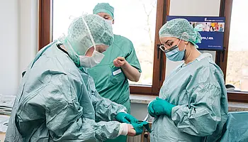 Mehrere Personen in OP-Kleidung demonstrieren Arbeitsschritte im Operationsbereich.