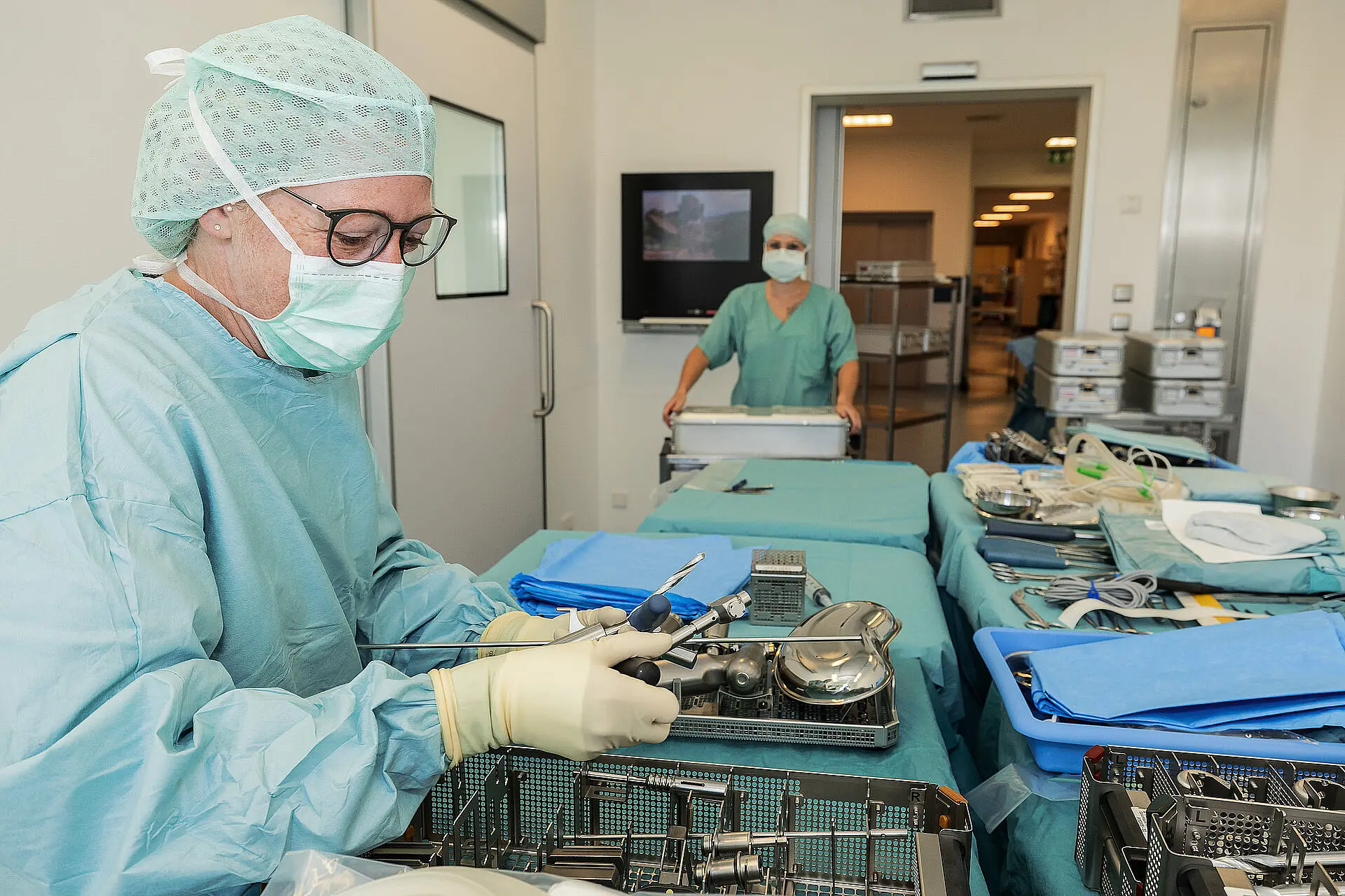 Eine Pflegekraft arbeitet konzentriert an einem Instrumententisch im Operationsbereich; im Hintergrund sind weitere OP-Arbeitsplätze sichtbar.