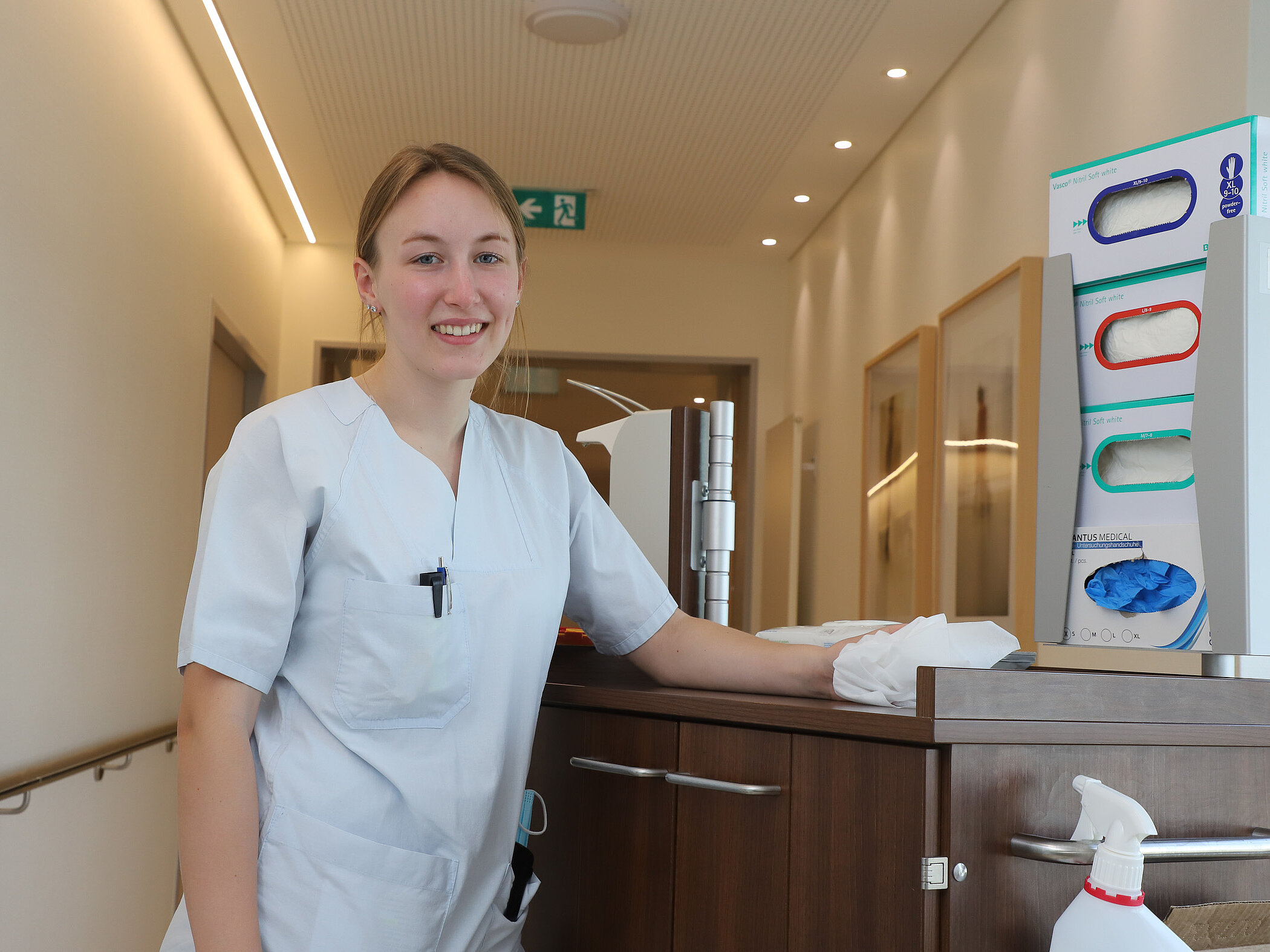 FSJlerin in heller Dienstkleidung steht lächelnd neben einem Pflegewagen auf einem Krankenhausflur.