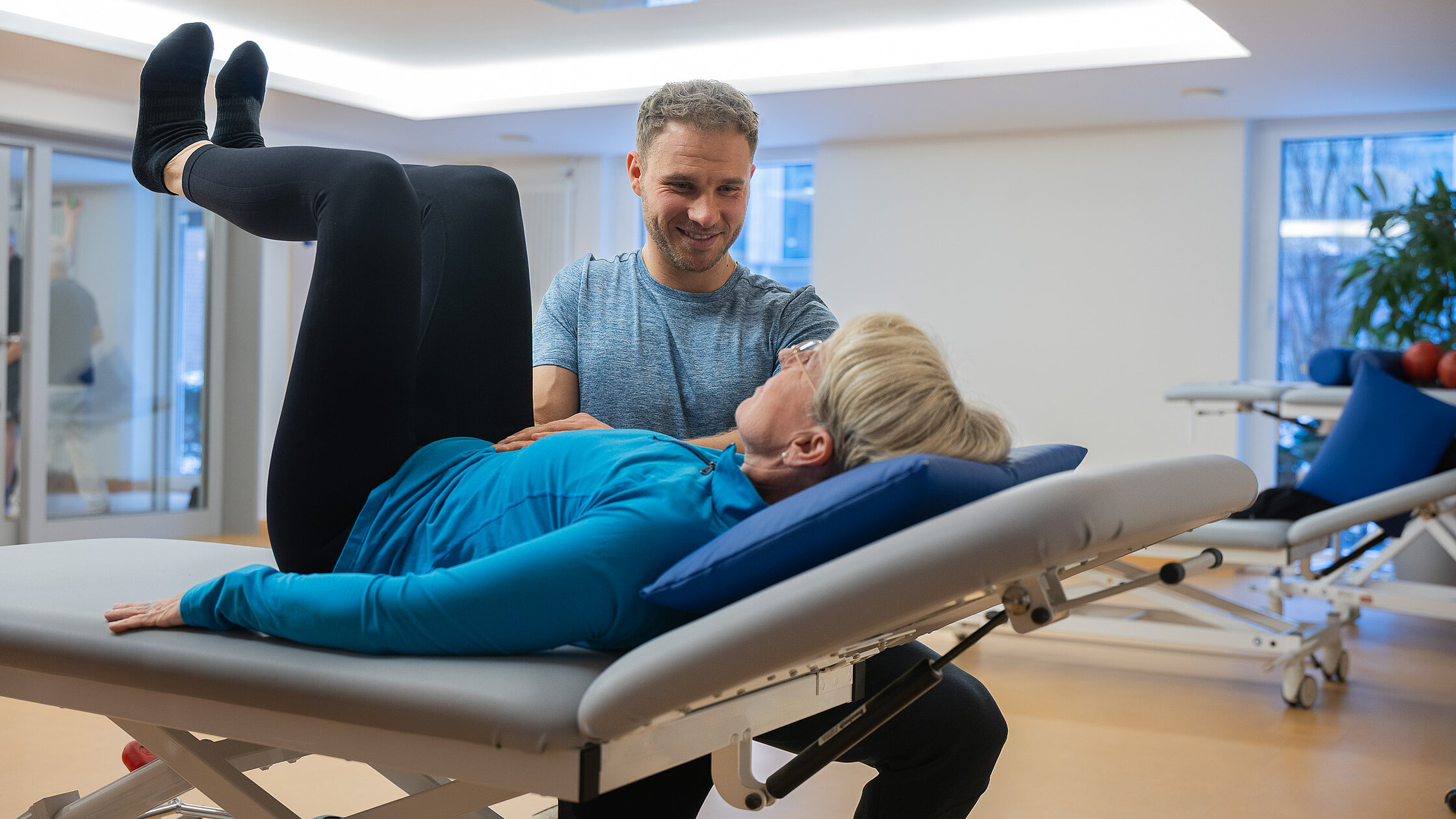 Eine Patientin liegt auf einer Therapieliege und führt eine Übung aus, während ein Therapeut sie anleitet.