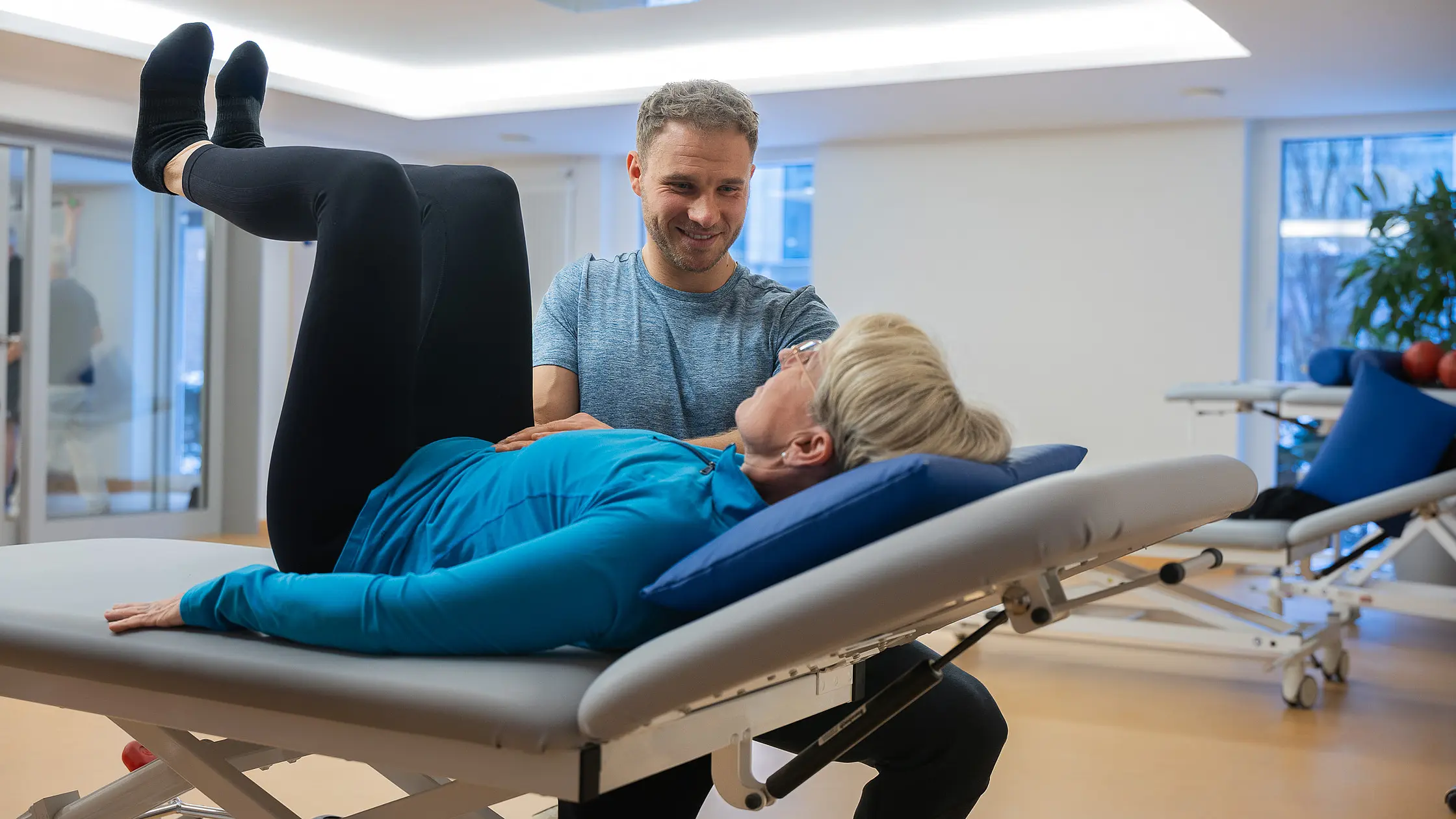 Eine Patientin liegt auf einer Therapieliege und führt eine Übung aus, während ein Therapeut sie anleitet.