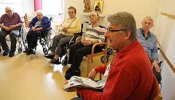 Mehrere Seniorinnen und Senioren sitzen im Stuhlkreis, während ein Mann mit einer Zeitung eine Gesprächsrunde moderiert.