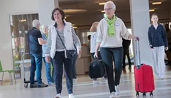 Zwei ehrenamtlich Engagierte mit grünem Halstuch gehen gemeinsam mit einer Besucherin durch den Eingangsbereich der Klinik.