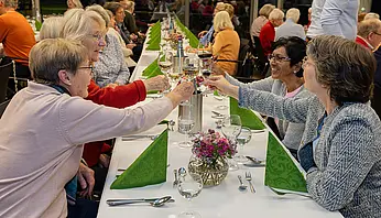 Mehrere Frauen sitzen an einer festlich gedeckten langen Tafel und stoßen mit Weingläsern an.